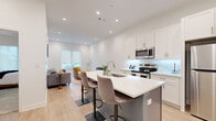 This thoughtfully designed single-floor residence offers a seamless blend of modern aesthetics and functional living. The open-concept layout connects the kitchen and living area, creating a bright, airy atmosphere enhanced by recessed lighting and light-toned hardwood flooring throughout.

The kitchen is a standout feature, showcasing crisp white shaker cabinetry with gold-toned hardware, a white quartz countertop island with seating for multiple bar-height chairs, stainless steel appliances including a refrigerator, range, and microwave, and a subway tile backsplash with subtle under-cabinet lighting. The entry door features a narrow sidelight window, welcoming natural light into the space.

The living area flows naturally from the kitchen, offering a versatile layout with room for comfortable seating arrangements and a dedicated nook bathed in natural light from roller-shaded windows.

The bedroom is generously proportioned, featuring plush carpeting, a large window with roller shades, and a dedicated workspace area. The room exudes a calm, neutral ambiance with tasteful wall art and warm lighting.

The bathroom is finished with a marble-look quartz vanity countertop, a framed mirror, and brushed nickel and gold accent fixtures, completing the residence's cohesive, contemporary design palette.