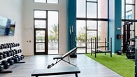 Located at Vintage Commerce in Fort Myers, Florida, this two-story fitness center is a well-appointed facility designed to support a full range of workout needs. The ground floor makes an immediate impression with its soaring double-height ceilings and floor-to-ceiling windows framed by teal accent columns, flooding the space with natural light and offering direct views of the outdoor pool and palm tree-lined surroundings. This level features a dedicated strength and functional training zone laid with artificial turf, equipped with Rogue power racks, barbell stations, a cable machine, weight storage, and adjustable benches. Hardwood-style flooring extends throughout the remaining ground floor areas, where additional free weights and open training space are available. A striking black spiral staircase connects the two levels, adding an architectural focal point to the interior. The upper floor offers a more intimate setting with a clean, well-lit cardio suite featuring multiple treadmills and other aerobic machines, all set on warm hardwood flooring with a large wall-mounted television. Glass railings along the mezzanine overlook the turf zone below, maintaining a sense of openness throughout. The overall ambiance is modern and energetic, combining bold design elements with functional, high-quality fitness equipment.