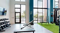 Located at Vintage Commerce in Fort Myers, Florida, this two-story fitness center is a well-appointed facility designed to support a full range of workout needs. The ground floor makes an immediate impression with its soaring double-height ceilings and floor-to-ceiling windows framed by teal accent columns, flooding the space with natural light and offering direct views of the outdoor pool and palm tree-lined surroundings. This level features a dedicated strength and functional training zone laid with artificial turf, equipped with Rogue power racks, barbell stations, a cable machine, weight storage, and adjustable benches. Hardwood-style flooring extends throughout the remaining ground floor areas, where additional free weights and open training space are available. A striking black spiral staircase connects the two levels, adding an architectural focal point to the interior. The upper floor offers a more intimate setting with a clean, well-lit cardio suite featuring multiple treadmills and other aerobic machines, all set on warm hardwood flooring with a large wall-mounted television. Glass railings along the mezzanine overlook the turf zone below, maintaining a sense of openness throughout. The overall ambiance is modern and energetic, combining bold design elements with functional, high-quality fitness equipment.