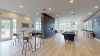 This single-floor clubhouse in Weatogue, CT is thoughtfully designed to provide a welcoming and versatile community gathering space. The open-concept layout is anchored by a striking dark tile fireplace column that serves as both a functional centerpiece and a bold architectural statement, dividing the space while maintaining a sense of openness throughout.

Light wood-tone flooring extends across the main recreation area, which features a full-size billiards table and adjustable bar-height tables with industrial-style stools, creating an ideal setting for social gatherings. Recessed lighting and pendant fixtures provide consistent, well-distributed illumination throughout.

A generously sized lounge area is carpeted and furnished with contemporary sofas and accent chairs in neutral and teal tones, complemented by a large wall of windows that frame views of the surrounding wooded landscape and outdoor pool. The natural light poured in through these oversized windows lends the space a bright, airy atmosphere.

The clubhouse also includes a well-appointed kitchen featuring white shaker cabinetry, dark countertops, a large island with a white surface, and a stainless steel sink — suitable for hosting events and community functions. Additional seating nooks with armchairs offer quieter, more intimate spaces for relaxation or conversation, all benefiting from the abundant natural light and scenic views that define this community facility.