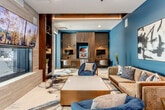 Located at 4401 Wornall Rd in Kansas City, Missouri, this sophisticated single-floor lounge space showcases a carefully curated contemporary interior design. The space is defined by a striking color palette, with bold teal accent walls contrasting against warm wood tones, dark cabinetry, and light-toned flooring. A standout marbled area rug anchors the main seating area, featuring fluid swirls of navy, gold, and cream that echo the artistic theme carried throughout the space.

The lounge area is furnished with plush velvet seating, including a taupe sofa and deep navy accent chairs arranged around a metallic gold-trimmed coffee table. Abstract artwork adorns the walls, contributing to the space's gallery-like atmosphere. A central media wall features a mounted flat-screen television flanked by dark built-in shelving units displaying decorative objects, with a fireplace insert below adding a focal point.

A dedicated business center alcove is integrated seamlessly into the design, equipped with workstations, task lighting, monitor displays, and overhead cabinetry in rich wood finishes. Geometric-patterned wallcovering in a separate nook adds textural depth. Recessed and linear LED lighting throughout enhances the modern, polished ambiance of the entire space.