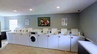 This single-floor laundry facility is designed for convenient, shared use. The room features a lineup of numbered washing machines and dryers arranged along one wall, providing ample capacity for multiple users. The machines are clearly labeled for easy identification, and "Easy Washing" instructional posters are mounted on either side of the room, offering guidance to users. A framed piece of wall art featuring a dog in a sports uniform adds a lighthearted, welcoming touch to the space. The walls are painted in a soft gray-green tone, creating a clean and neutral atmosphere. Recessed ceiling lights provide consistent, even illumination throughout the room, and the textured ceiling contributes to the practical, utilitarian character of the space. The flooring appears to be durable and easy to maintain, well-suited for a high-traffic laundry environment. Overall, the facility is thoughtfully organized and maintained, offering a functional and approachable space for laundry needs.