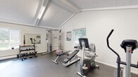 This single-floor fitness center is designed to support a comprehensive workout experience in a bright, welcoming environment. The space is defined by its striking vaulted ceilings with exposed white beams, which lend an open, airy feel throughout. Fluorescent lighting supplements the abundant natural light that streams in through large sliding glass doors and multiple windows, offering views of a surrounding outdoor pool area and lush greenery.

The facility is divided into two connected workout zones. The cardio area features treadmills, an elliptical trainer, a stationary recumbent bike, and a stair climber, all arranged on durable rubber flooring. Large floor-to-ceiling sliding doors in this section provide a seamless visual connection to the outdoors, making workouts feel expansive and energizing.

The strength training area is equally well-appointed, housing a multi-functional cable machine, an adjustable workout bench, a dumbbell rack, and a stability ball. Wall-mounted mirrors throughout both zones enhance the sense of space and allow users to monitor their form during exercise. A wall-mounted air conditioning unit ensures year-round comfort. The clean white walls, textured ceilings, and dark rubber flooring create a functional yet polished aesthetic that is both practical and motivating.