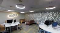 This contemporary workspace combines functional design with distinctive aesthetic elements to create an engaging professional environment. The most striking feature is a full accent wall adorned with three-dimensional sculptural panels, their organic, pebble-like relief texture adding depth and visual interest to the room. The ceiling is outfitted with a series of circular ring pendant lights that cast a bright, even illumination throughout the space, complemented by circular wall-mounted light fixtures that reinforce the cohesive design language.

The flooring consists of modular carpet tiles in a geometric pattern with grey and yellow tones, adding warmth and visual texture underfoot. Workstations featuring rounded white desks are paired with ergonomic task chairs in grey and yellow, promoting both comfort and productivity. A dedicated lounge area offers a brown upholstered armchair and a two-seat sofa in a complementary warm tone, providing a more relaxed setting for informal meetings or breakout sessions. The overall color palette—neutral greys accented with gold and yellow tones—creates a professional yet inviting ambiance suited to a modern office or collaborative workspace.
