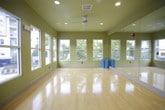 This single-floor fitness and yoga studio is thoughtfully designed to support a wide range of exercise and wellness activities. The open floor plan is anchored by gleaming, light-toned hardwood floors that reflect natural light and create a clean, energizing atmosphere throughout the space.

A row of large, tall windows lines one wall, flooding the interior with abundant natural light while offering views of outdoor amenity areas, mature trees, and the surrounding residential community. The warm olive-green walls lend a calming, grounded tone to the environment, complementing the bright flooring and white trim.

Full-length mirrors are positioned along one wall, a practical and essential feature for fitness and movement-based activities, visually expanding the room while allowing participants to monitor their form. Rolled yoga mats stored along the base of the mirror wall signal the studio's primary use.

The studio is equipped with a wall-mounted flat-screen television, ideal for guided workout sessions or instructional content. A ceiling fan with integrated recessed lighting ensures proper ventilation and consistent illumination. Multiple access points, including glass-paneled doors, provide easy entry and exit, and the layout flows openly to accommodate group classes or individual training sessions. The overall ambiance is clean, functional, and welcoming.