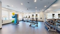 Located at 3360 Chichester Ave in Boothwyn, PA, this fitness center offers a thoughtfully designed exercise space across a single floor. The room is finished with durable rubber flooring and bright white walls accented by a cool blue-gray feature wall, creating a clean and energizing atmosphere. Recessed lighting and a ceiling fan ensure the space remains well-lit and comfortable during workouts.

Natural light floods the room through multiple large windows fitted with roller shades, offering views of the surrounding outdoor landscape. The main exercise area houses a row of treadmills and elliptical trainers, each equipped with individual monitors, as well as a stationary spin bike. A dedicated strength training section includes a cable/functional trainer machine, a weight bench, and a full dumbbell rack displayed in front of a large wall-mounted mirror — ideal for form monitoring and making the space feel more expansive.

Complementing the cardio and strength equipment is a colorful medicine ball storage rack, adding a functional yet vibrant element to the room. The layout is open and spacious, allowing for easy movement between stations. Glass-paneled interior doors connect to adjacent areas, enhancing the sense of connectivity. Overall, this fitness center is a comprehensive and inviting facility well-suited for a wide range of workout routines.