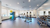 Located at 3360 Chichester Ave in Boothwyn, PA, this fitness center offers a thoughtfully designed exercise space across a single floor. The room is finished with durable rubber flooring and bright white walls accented by a cool blue-gray feature wall, creating a clean and energizing atmosphere. Recessed lighting and a ceiling fan ensure the space remains well-lit and comfortable during workouts.

Natural light floods the room through multiple large windows fitted with roller shades, offering views of the surrounding outdoor landscape. The main exercise area houses a row of treadmills and elliptical trainers, each equipped with individual monitors, as well as a stationary spin bike. A dedicated strength training section includes a cable/functional trainer machine, a weight bench, and a full dumbbell rack displayed in front of a large wall-mounted mirror — ideal for form monitoring and making the space feel more expansive.

Complementing the cardio and strength equipment is a colorful medicine ball storage rack, adding a functional yet vibrant element to the room. The layout is open and spacious, allowing for easy movement between stations. Glass-paneled interior doors connect to adjacent areas, enhancing the sense of connectivity. Overall, this fitness center is a comprehensive and inviting facility well-suited for a wide range of workout routines.