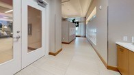 This single-floor lobby and mail room area presents a sleek, contemporary aesthetic designed to make a strong first impression. Large-format light-toned tile flooring runs throughout, lending a clean and polished look that unifies the space. Recessed ceiling lighting provides even, well-distributed illumination across all areas.

The mail room corridor is a standout feature, with rows of white built-in U.S. Mail mailboxes neatly mounted along the walls, complemented by warm wood baseboards that add a touch of natural warmth to the otherwise neutral palette. Sheer curtains frame a window at the far end of the corridor, allowing soft natural light to filter in.

An adjacent hallway showcases dramatic full-wall graphic murals with a geometric, architectural pattern rendered in earth tones and deep contrasts, creating a bold visual focal point. This corridor leads to a secure exit door, with the murals continuing on both sides for an immersive effect.

A service counter area features warm wood cabinetry beneath a white countertop, with a wall-mounted flat-screen TV displaying news content above it. A small potted plant adds a touch of greenery. Throughout the space, the design balances functionality with refined style, using a neutral color palette of warm whites, tans, and wood accents to create an inviting and professional environment.