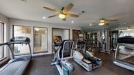 Located at 501 Napa Valley Dr in Little Rock, Arkansas, this single-floor fitness center is a well-equipped exercise facility designed to support a comprehensive workout routine. The space is outfitted with a selection of True-brand cardio machines, including treadmills, ellipticals, and stationary bikes, providing residents with a variety of aerobic training options. A dedicated strength training area features a multi-station cable machine, with floor-to-ceiling mirrored walls that visually expand the space and allow users to monitor their form during exercise. The rubber flooring throughout provides both durability and safety underfoot. Large windows line one side of the facility, inviting generous amounts of natural light and offering views of the surrounding landscaped grounds and brick residential buildings. Ceiling fans are installed throughout, contributing to air circulation and comfort during workouts. Recessed lighting supplements natural light, ensuring the space remains well-lit at all times. A framed exercise chart is posted on the wall, serving as a practical reference for users. The neutral gray walls and clean, functional layout create an environment that is organized and focused, making this fitness center a practical and accessible amenity for those at this Little Rock address.