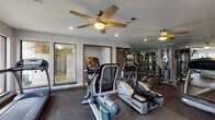 Located at 501 Napa Valley Dr in Little Rock, Arkansas, this single-floor fitness center is a well-equipped exercise facility designed to support a comprehensive workout routine. The space is outfitted with a selection of True-brand cardio machines, including treadmills, ellipticals, and stationary bikes, providing residents with a variety of aerobic training options. A dedicated strength training area features a multi-station cable machine, with floor-to-ceiling mirrored walls that visually expand the space and allow users to monitor their form during exercise. The rubber flooring throughout provides both durability and safety underfoot. Large windows line one side of the facility, inviting generous amounts of natural light and offering views of the surrounding landscaped grounds and brick residential buildings. Ceiling fans are installed throughout, contributing to air circulation and comfort during workouts. Recessed lighting supplements natural light, ensuring the space remains well-lit at all times. A framed exercise chart is posted on the wall, serving as a practical reference for users. The neutral gray walls and clean, functional layout create an environment that is organized and focused, making this fitness center a practical and accessible amenity for those at this Little Rock address.