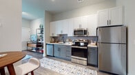 This thoughtfully designed single-floor studio offers a seamless open-concept layout that integrates the kitchen, living, and sleeping areas into one cohesive space. The kitchen is a standout feature, showcasing a two-tone cabinet design with crisp white upper cabinets and slate-gray lower cabinets, complemented by stainless steel appliances including a refrigerator, electric range, over-the-range microwave, and dishwasher. White quartz countertops and a matte black faucet add a polished, modern aesthetic, while wood-look plank flooring flows consistently throughout the unit. The main living and sleeping area is bright and welcoming, highlighted by a bold teal accent wall that adds depth and character. Large windows invite natural light and offer views of the surrounding greenery, enhancing the open feel of the space. A walk-in closet provides generous storage directly off the main room. The bathroom is finished in a clean, contemporary style featuring a floating white vanity with black hardware, wood-look tile flooring, and neutral gray walls. Every detail of this studio reflects a modern design sensibility, creating a functional and visually cohesive living environment.