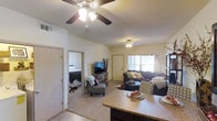 This single-floor residential unit offers a thoughtfully designed open-concept layout that seamlessly connects the kitchen, dining, and living areas. The kitchen features a dark-toned island with granite-style countertops, providing both functional workspace and a natural divide between the cooking and living spaces. The living room is warm and inviting, centered around a flat-screen TV and comfortable sectional seating, with a ceiling fan overhead ensuring year-round comfort. A large window near the front entrance allows natural light to fill the space, enhancing its airy and welcoming ambiance. Neutral-toned carpet throughout the living and bedroom areas adds warmth underfoot. The bedroom, visible through an open doorway, is spacious and well-lit, featuring a neatly made bed with a bold patterned comforter, a ceiling-mounted light fixture, and its own area rug. The unit's layout promotes a sense of openness while maintaining defined areas for living, dining, and rest. Overall, the space presents a practical and comfortable interior well-suited for everyday living.