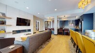 This stylish single-floor amenity space is designed with an open-concept layout that seamlessly connects a living area, dining zone, and full kitchen. The living area is anchored by a sleek, wall-mounted electric fireplace flanked by floating shelves, a wall-mounted television, and large windows that invite natural light and provide views of an outdoor terrace. Light wood-look flooring runs throughout, enhancing the bright and welcoming atmosphere.

The kitchen is a standout feature, showcasing deep navy cabinetry paired with white quartz countertops, a mosaic tile backsplash, stainless steel appliances including a built-in oven and dishwasher, and a stainless steel range hood. A spacious island with bar-height seating accommodates multiple guests, making it ideal for entertaining and communal gatherings.

Pendant lights with sculptural pleated shades hang above the island and dining counter, adding a refined decorative touch. The dining area offers ample seating with upholstered bar stools in rich brown tones and bold yellow accent chairs that inject energy and personality into the space. Recessed lighting throughout ensures a consistently well-lit environment. The overall design balances modern sophistication with functional, community-oriented comfort.