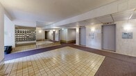 This residential building's common area lobby and hallway space occupies a generously sized single floor, offering a functional and well-organized entry experience for residents. The layout comprises two interconnected hallway sections that guide visitors through the space with clear sightlines and practical flow.

The main lobby area is finished with cream-toned square ceramic floor tiles, which transition into plush carpeted corridors leading toward the building's interior. A standout feature of the space is a striking decorative wall constructed of textured glass blocks, which spans a substantial portion of one wall and is further amplified by a full-length mirror panel beside it, creating an impressive visual depth and adding brightness to the interior.

Two elevator doors are prominently positioned along one wall, providing convenient vertical access throughout the building. Adjacent to the entry area, a large built-in mailbox unit accommodates the full complement of building residents, offering individual locked compartments in a neatly organized grid arrangement. An intercom panel is also visible, supporting secure building access.

The ceilings are finished with a textured surface, and wall-mounted sconce lighting provides warm, even illumination throughout. Baseboard heating units line the lower walls of the hallways, ensuring comfort during colder months. Overall, the space conveys a practical, no-frills design with durable, easy-to-maintain finishes suited to a high-traffic residential common area.