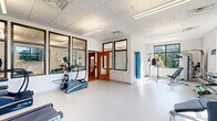 Located at 2800 Lawndale Dr in Greensboro, NC, this single-floor wellness facility is thoughtfully designed to support fitness and rehabilitation needs. The space is divided into two distinct areas that work in harmony to create a complete wellness environment.

The exercise room features durable, easy-to-clean flooring and is outfitted with a variety of fitness equipment, including treadmills, an elliptical machine, a cable weight machine, and stability balls. Bright, large windows allow natural light to fill the room, creating an open and energizing atmosphere. A wall-mounted display screen adds a modern touch, while the clean white walls and drop-panel ceiling keep the aesthetic clinical yet welcoming.

Adjacent to the exercise area is a stylishly appointed lounge space with warm hardwood flooring. Comfortable seating — including a cream sofa and plush teal accent chairs — is arranged around a rounded wood coffee table, creating an inviting area for relaxation or consultation. Large interior glass windows between the two rooms provide visual connectivity while maintaining distinct functional zones. The lounge also benefits from natural light through exterior windows.

Together, the two rooms form a cohesive, professional facility well-suited for physical therapy, rehabilitation, or community fitness programs.