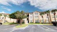 This single-floor residence is situated within a well-maintained multi-story building that reflects a classic Florida architectural style. The exterior is finished in a warm beige stucco, complemented by white-trimmed windows that allow natural light to filter throughout. The building features covered stairways providing access to upper-level units, along with open balconies that contribute to the structure's airy, coastal character.

The grounds surrounding the building are thoughtfully landscaped, featuring a mix of mature shade trees and tall palm trees that reinforce the tropical atmosphere. Neatly manicured lawns, decorative shrubs, and mulched planting beds line the building's foundation, creating a polished and inviting exterior appearance. A paved parking lot with designated accessible spaces sits directly in front of the building, offering straightforward access for residents and visitors alike.

The unit itself occupies a single floor within the building, offering a practical and comfortable layout. While interior images were not clearly available, the building's design and construction suggest a well-organized residential space suited for everyday living. The overall environment reflects a community-oriented Florida residential setting under bright, open skies.