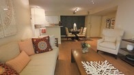This single-floor residence features an open-concept layout that seamlessly connects the living, dining, and kitchen areas, creating a cohesive and functional flow throughout the space. The living area is anchored by a light-toned sofa dressed with a vibrant, patterned throw pillow, paired with a warm wood coffee table adorned with decorative coral accents and a potted succulent, adding a natural touch to the interior. Hardwood flooring extends throughout the main living spaces, contributing warmth and continuity. A large floor lamp provides ambient lighting, complementing the track lighting overhead. The dining area, positioned just beyond the living space, features a round table surrounded by cushioned chairs with a bold, dark patterned accent wall serving as a dramatic backdrop — a clear design focal point. The kitchen is visible from the living area, outfitted with white cabinetry, white appliances, and a raised bar-style counter that separates it from the dining space. The entry door is visible along the far wall, reinforcing the compact yet efficient single-floor layout. Overall, the space presents a warm, inviting ambiance with an eclectic mix of textures and décor.