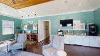 Located at 531 Old Maccumber Station Dr in Wilmington, NC, this single-story community space is thoughtfully designed with a vibrant coastal aesthetic. The interior is defined by rich teal accent walls, crisp white trim, and gleaming polished concrete floors that run throughout, creating a cohesive and modern atmosphere.

The main gathering area features a tufted leather sofa flanked by woven side tables and table lamps, set against large windows framed by floor-length navy drapes that overlook a community pool area. Multiple seating arrangements, including upholstered lounge chairs and a casual high-top dining table with modern white chairs, provide flexible spaces for lounging or casual meetings.

A dedicated workstation area is equipped with computer monitors and wooden desks, supported by pendant lighting with warm rattan shades. A wall-mounted flat-screen TV and a service counter with granite countertops, a printer, and a compact refrigerator add functional convenience. Adjacent hallways connect to additional private rooms, suggesting offices or restrooms.

Vaulted ceilings with natural wood paneling and clerestory windows in the main area amplify the sense of height and airiness, while abundant windows throughout flood the space with natural light and offer views of the surrounding outdoor amenities. The overall ambiance is welcoming, functional, and distinctly coastal in character.