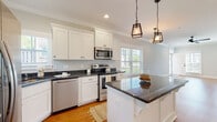 This two-story home is designed with a clean, contemporary aesthetic and a neutral color palette throughout. The main level centers on an open-concept layout where the living area flows directly into the kitchen. Hardwood flooring extends across the main level, complemented by crown molding and recessed lighting. A staircase with white balusters and warm wood treads connects both floors, accompanied by a ceiling fan that anchors the living space.

The kitchen is a standout feature, outfitted with white shaker-style cabinetry, dark granite countertops, and stainless steel appliances including a refrigerator, range, microwave, and dishwasher. A central island provides additional prep space and casual seating, while pendant lantern-style light fixtures add character. Multiple windows allow natural light to fill the space. A compact powder room with a pedestal sink and hardwood flooring is also located on the main level.

Upstairs, bedrooms feature plush carpeting, crown molding, ceiling fans, and generous closet space, including walk-in configurations. Bathrooms on the upper level include white vanities, tile flooring, and tub-shower combinations, maintaining the home's cohesive, neutral design throughout.