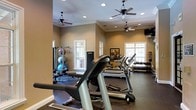 This dedicated fitness center offers a comprehensive workout environment designed to accommodate a wide range of exercise routines. The space is finished with warm, neutral-toned walls and crowned with elegant crown molding, lending it a polished, refined atmosphere uncommon in typical gym settings. Multiple ceiling fans are evenly distributed throughout, ensuring comfortable air circulation during workouts, while recessed lighting provides consistent, bright illumination across the entire floor.

Natural light pours in through several white-framed windows along the walls, creating an open and inviting ambiance. The flooring is a dark, durable gym-appropriate surface well-suited for heavy equipment and high-traffic use.

The cardio section is prominently featured, with multiple treadmills arranged to allow ample space between users. Along one wall, a dedicated free weights area hosts a full rack of dumbbells spanning a wide range of sizes, ideal for strength training. A multi-station cable and weight machine anchors another corner, accompanied by a selection of medicine and stability balls for functional fitness exercises. A wall-mounted flat-screen television provides entertainment for users during their workouts. The overall layout is thoughtfully organized, balancing equipment density with open floor space to ensure a safe and efficient exercise experience.