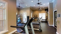 This dedicated fitness center offers a comprehensive workout environment designed to accommodate a wide range of exercise routines. The space is finished with warm, neutral-toned walls and crowned with elegant crown molding, lending it a polished, refined atmosphere uncommon in typical gym settings. Multiple ceiling fans are evenly distributed throughout, ensuring comfortable air circulation during workouts, while recessed lighting provides consistent, bright illumination across the entire floor.

Natural light pours in through several white-framed windows along the walls, creating an open and inviting ambiance. The flooring is a dark, durable gym-appropriate surface well-suited for heavy equipment and high-traffic use.

The cardio section is prominently featured, with multiple treadmills arranged to allow ample space between users. Along one wall, a dedicated free weights area hosts a full rack of dumbbells spanning a wide range of sizes, ideal for strength training. A multi-station cable and weight machine anchors another corner, accompanied by a selection of medicine and stability balls for functional fitness exercises. A wall-mounted flat-screen television provides entertainment for users during their workouts. The overall layout is thoughtfully organized, balancing equipment density with open floor space to ensure a safe and efficient exercise experience.