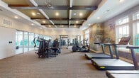 This fitness center is designed to offer a comprehensive workout experience across two distinct zones. The main exercise floor is finished with durable carpet and features an extensive selection of strength training machines and cardio equipment, including treadmills, ellipticals, and cable machines, as well as a medicine ball rack. A full-length mirror along one wall visually expands the space and aids in form correction during workouts. The ceiling is a notable architectural highlight, showcasing a coffered design with dark exposed wooden beams, recessed lighting, and a ceiling fan, contributing to a refined yet functional ambiance. A textured, earth-toned accent wall adds an artistic focal point near the cardio equipment. Large windows allow ample natural light to fill the space while providing outdoor views. A separate studio room, enclosed by glass-paneled walls and flooded with natural light through a bank of oversized windows, offers a clean, open floor with specialized equipment, making it well-suited for stretching, yoga, or group training sessions. Access to an adjacent common area is available through glass-paned doors.