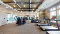 This fitness center is designed to offer a comprehensive workout experience across two distinct zones. The main exercise floor is finished with durable carpet and features an extensive selection of strength training machines and cardio equipment, including treadmills, ellipticals, and cable machines, as well as a medicine ball rack. A full-length mirror along one wall visually expands the space and aids in form correction during workouts. The ceiling is a notable architectural highlight, showcasing a coffered design with dark exposed wooden beams, recessed lighting, and a ceiling fan, contributing to a refined yet functional ambiance. A textured, earth-toned accent wall adds an artistic focal point near the cardio equipment. Large windows allow ample natural light to fill the space while providing outdoor views. A separate studio room, enclosed by glass-paneled walls and flooded with natural light through a bank of oversized windows, offers a clean, open floor with specialized equipment, making it well-suited for stretching, yoga, or group training sessions. Access to an adjacent common area is available through glass-paned doors.