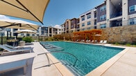 This expansive outdoor amenity area centers around a generously sized swimming pool with crystal-clear turquoise water, designed to provide a resort-like experience. The pool deck is finished in light-toned pavers and features built-in in-water lounge chairs that allow residents to relax partially submerged, ideal for warm sunny days. A striking wood-framed pergola anchors one side of the pool, offering shaded seating beneath its open-beam canopy and creating a comfortable gathering point. On the opposite side, large cantilever shade umbrellas are strategically positioned over rows of cushioned lounge chairs, providing ample sun protection across the deck. The entire area is enclosed by a black iron fence, ensuring both security and an open, airy feel. A substantial natural limestone retaining wall runs along one edge of the property, adding texture and a distinctive architectural character to the outdoor space. Thoughtfully maintained landscaping with ornamental grasses, native shrubs, and mulched planting beds softens the hardscape and contributes to the overall aesthetic. The multi-story residential buildings surrounding the pool complete the backdrop, blending stone masonry and contemporary architectural design. Under open skies, this outdoor area offers a bright, welcoming, and well-appointed environment for relaxation and recreation.
