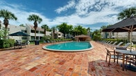 Located at 15690 Bull Run Rd in Miami Lakes, Florida, this expansive outdoor amenity area offers a resort-style experience defined by thoughtful design and lush South Florida surroundings. At the heart of the space is a generously sized oval swimming pool with crystal-clear blue water, reaching a depth of 6 feet and framed by smooth curved coping. The pool deck is finished in warm terracotta tiles that extend broadly around the entire perimeter, providing ample room for relaxation and recreation. Rows of contemporary lounge chairs are arranged along multiple sides of the pool, offering abundant sunbathing space for residents and guests. Towering palm trees are strategically placed throughout the deck, delivering natural shade and reinforcing the tropical ambiance. Manicured hedgerows and raised planter beds with sculpted shrubs define the boundaries of the space, adding structure and greenery. A covered pavilion-style structure with a clay tile roof anchors one end of the complex, housing support amenities and providing a shaded gathering area. The surrounding multi-story buildings, clad in neutral stucco with red tile rooflines and private balconies, frame the pool area and contribute to the cohesive Mediterranean-inspired architectural character of the community. Open skies and abundant natural light complete the inviting outdoor atmosphere.