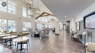 Located at 152 Market St in Pittsboro, NC, this impressive single-floor community clubhouse offers a generously proportioned, amenity-rich environment designed for comfort and connection. The space is defined by soaring vaulted ceilings with exposed wooden beams, crisp white walls, and durable wood-look flooring that flows seamlessly throughout.

The main living areas are open and airy, bathed in natural light from expansive double-hung windows that frame views of an outdoor pool and surrounding grounds. Dramatic geometric chandelier pendants illuminate the gathering spaces, which feature a mix of plush seating arrangements, a statement fireplace, and a mounted television for entertainment.

A dedicated game room offers a full-size billiards table, shuffleboard, and casual seating, while a stylish café area provides counter seating, café-style tables, and a self-serve refreshment station with refrigerated beverages and coffee service. Built-in shelving and gray wainscoting add refined architectural detail throughout.

Connecting hallways lead to a well-equipped fitness center, visible through sleek glass-paneled walls, and a dedicated Health & Wellness suite accessed via a frosted-glass door. The overall ambiance is modern yet welcoming, blending contemporary design with warm, residential touches suited to an active community lifestyle.