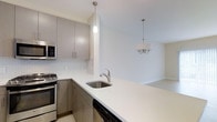 This three-floor residence at 9C showcases a clean, contemporary design highlighted by a well-appointed kitchen that serves as a central feature of the living space. The kitchen is outfitted with sleek, taupe-toned cabinetry equipped with brushed nickel hardware, offering ample storage through both upper and lower cabinets. Stainless steel appliances, including a gas range, built-in microwave, and dishwasher, complement the modern aesthetic. A white quartz countertop extends into a peninsula, providing additional workspace and a natural transition into the adjoining living and dining areas. A white mosaic tile backsplash adds subtle texture to the kitchen walls. Pendant lighting above the peninsula and a chandelier over the dining area provide layered illumination throughout the open-plan space. The neutral color palette of soft whites and warm grays carries throughout, contributing to a bright and airy atmosphere. The layout across three floors allows for distinct separation of living, sleeping, and functional spaces, offering both versatility and privacy within the residence.
