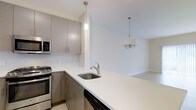 This three-floor residence at 9C showcases a clean, contemporary design highlighted by a well-appointed kitchen that serves as a central feature of the living space. The kitchen is outfitted with sleek, taupe-toned cabinetry equipped with brushed nickel hardware, offering ample storage through both upper and lower cabinets. Stainless steel appliances, including a gas range, built-in microwave, and dishwasher, complement the modern aesthetic. A white quartz countertop extends into a peninsula, providing additional workspace and a natural transition into the adjoining living and dining areas. A white mosaic tile backsplash adds subtle texture to the kitchen walls. Pendant lighting above the peninsula and a chandelier over the dining area provide layered illumination throughout the open-plan space. The neutral color palette of soft whites and warm grays carries throughout, contributing to a bright and airy atmosphere. The layout across three floors allows for distinct separation of living, sleeping, and functional spaces, offering both versatility and privacy within the residence.