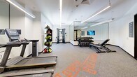 Located at 1600 Broadway in Sacramento, California, this fitness center offers a thoughtfully designed exercise space for a well-rounded workout experience. The room features a clean, modern aesthetic with crisp white walls, recessed linear LED lighting, and durable speckled carpet flooring throughout. Bold orange graphic lettering on the floor adds a dynamic, energetic visual element to the space.

The facility is equipped with a comprehensive selection of fitness equipment, including Matrix treadmills and an elliptical machine, each paired with individual monitor screens for interactive cardio sessions. A multi-functional cable trainer anchors the strength training zone, while a full rack of graduated dumbbells and adjustable benches provide ample free-weight options. A medicine ball and accessory storage rack rounds out the functional training area.

A floor-to-ceiling mirror wall on one side of the room visually expands the space and supports proper form during workouts, with a wall-mounted flat-screen TV integrated into the mirror zone. Natural light filters in through windows offering views of surrounding neighborhood greenery. The entrance features glass-paned doors and includes a water fountain station for convenience. Informational signage near the entrance provides facility guidelines, contributing to an organized and user-friendly environment.