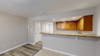 This single-floor space is thoughtfully laid out to maximize functionality and flow. The open-concept design connects the kitchen and living area seamlessly, creating a cohesive and practical environment. Neutral gray walls and white trim throughout lend a clean, consistent aesthetic, while the transition between light tile flooring in the kitchen zone and wood-look laminate in the living area subtly defines each space.

The kitchen is equipped with warm honey-toned wood cabinetry offering generous upper storage, granite-look laminate countertops, a double basin sink, a white electric range, and a black refrigerator. Recessed lighting illuminates the workspace effectively. A peninsula counter extends from the kitchen into the living area, featuring bracket-supported overhang seating — ideal for casual dining or meal prep. Additional upper cabinets line the peninsula wall, providing further storage.

The living area is open and versatile, offering a generous footprint with wood-look laminate flooring, a textured ceiling, and access to the entry door with an adjacent mirrored closet. A wall-mounted intercom panel is also present near the entry.

The bathroom is finished in clean whites, featuring tile flooring, a single-sink vanity with wood cabinetry, a toilet, and a full-size soaking tub with shower surround.