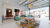 Located at 150 N 3rd St in the heart of downtown Columbus, Ohio, this single-floor amenity space blends modern industrial design with a lively, social atmosphere. The open-plan layout is anchored by large-format tile flooring, soaring ceilings with exposed ductwork, and structural columns that add an urban, loft-inspired character throughout.

The main lounge area is generously proportioned, featuring a full-size pool table set atop a bold geometric-patterned area rug, surrounded by eclectic seating arrangements in teal, orange, and green upholstery. Pendant lighting and wall-mounted televisions complete the social atmosphere, while exposed brick accent panels add a warm, textural contrast to the crisp white walls.

A fully equipped kitchen anchors one side of the space, outfitted with stainless steel appliances, warm wood cabinetry, a dark herringbone tile backsplash, and an extended island with white countertops and bar seating beneath striking cage-style pendant lights — ideal for communal gatherings.

Floor-to-ceiling glass garage doors open the space directly to an outdoor terrace, flooding the interior with natural light and providing panoramic views of the Columbus skyline. Additional seating zones throughout the space offer flexible arrangements for both relaxed socializing and more intimate gatherings, creating a dynamic and well-appointed community environment.