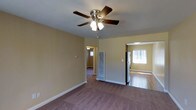This single-floor residence offers a practical and comfortable layout with a warm, neutral color palette throughout. The main living area features plush carpeting underfoot and soft yellow-toned walls that create an inviting atmosphere. A ceiling fan with an integrated light fixture serves as a central element, providing both ambient lighting and air circulation. White baseboards provide a clean contrast against the carpeted flooring and painted walls.

The open sightlines connect the living area to the adjoining kitchen, which transitions to hardwood flooring, offering a clear distinction between the two spaces. The kitchen is equipped with a stainless steel refrigerator and benefits from a window that allows natural light to brighten the area. A floor-standing radiator unit is positioned between the living and kitchen spaces, serving as the primary heat source.

A hallway visible from the main living area leads further into the home, hinting at additional rooms beyond. The hallway is lit with a ceiling-mounted fixture and continues the warm wall tones seen throughout. The overall layout feels connected and functional, with a straightforward flow between spaces. The design is understated and practical, making it well-suited for everyday living.