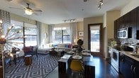 This single-floor space showcases a thoughtfully designed open-concept layout that effortlessly blends the dining and living areas into one cohesive, functional environment. The dining area features a sleek glass-top table accompanied by a mix of modern chairs, including a standout yellow accent chair that adds a vibrant pop of color. Adjacent to the dining space, the living area is anchored by a generous sectional sofa dressed with colorful throw pillows, complemented by a bold geometric-patterned area rug in blue and white tones that defines the lounge zone.

Natural light floods the interior through large windows dressed with patterned curtains, creating a bright and welcoming atmosphere. Track lighting installed along the ceiling provides additional illumination and highlights the room's contemporary character. Dark-finished flooring runs throughout, offering a striking contrast against the neutral wall tones and lighter furnishings.

A glass door with a security screen opens to the outdoors, offering a view of the surrounding greenery and extending the sense of openness. The ceiling fan adds a practical element while contributing to the relaxed, modern ambiance. Overall, the space presents a well-balanced blend of style, comfort, and functionality.