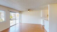 This single-floor unit offers a clean, functional layout with consistent wood-style flooring running throughout the living areas and bedrooms, creating a cohesive and easy-to-maintain interior. The open living and dining area is generously sized, featuring a ceiling fan with integrated light fixture and a textured ceiling. A sliding glass door floods the space with natural light and provides direct access to an outdoor balcony, while a secondary window further enhances the bright, airy atmosphere.

The kitchen is designed in a galley configuration, outfitted with white cabinetry extending to the ceiling for ample storage. Appliances include a gas cooktop, wall oven, and over-the-range microwave. A pass-through counter connects the kitchen to the living area, supporting an open feel.

The bedrooms each feature wood-style flooring, ceiling fans, and natural light from windows fitted with blinds or vertical slat coverings. One bedroom includes a sliding closet with a full-width door panel. The bathroom presents a clean, all-white finish with a shower-tub combination, a window above the tub for ventilation, a wall-mounted mirror, and a single-basin vanity.