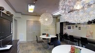 This single-floor space combines creative design elements with a functional, welcoming layout. The room is defined by its eye-catching lighting fixtures — large, ornate white dandelion-style pendant lights hang from the ceiling, creating a bold artistic focal point that immediately draws the eye upward. Track lighting and recessed fluorescent fixtures supplement the pendants, ensuring the space is well-lit throughout.

The flooring consists of slate tile in earthy, muted tones, lending a grounded, textured quality to the room. White round tables are paired with sleek black chairs, arranged to accommodate multiple groups simultaneously, suggesting a communal or multi-use purpose.

A standout feature is the wall typography, where an inspirational quote — referencing setting the world "on fire" — is displayed prominently in dark lettering against a warm beige wall, adding a motivational and expressive character to the space. Built-in cabinetry and shelving along one wall provide storage and display space, while framed artwork and colorful accents contribute to a lively, creative atmosphere. The double-panel door offers a clean, traditional contrast to the room's more eclectic decorative elements.