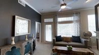 This two-story residence offers a thoughtfully designed interior that balances comfort and contemporary style. The living area serves as a welcoming focal point, featuring large windows dressed with layered blinds and patterned curtains that allow generous amounts of natural light to fill the space. A ceiling fan overhead provides both function and a finishing architectural touch, while crown molding along the ceiling adds a refined, classic detail. The walls are finished in a rich slate-blue accent color that contrasts elegantly with the neutral taupe tones found throughout, creating a warm yet modern ambiance. Soft carpeted flooring underfoot contributes to the cozy atmosphere of the main living space. A glass-paneled exterior door provides access to the outdoors, enhancing the sense of openness and connectivity to the outside. Decorative touches, including a rustic wooden console, mirror accents, and layered textiles, complement the architectural features and give the space a curated, lived-in feel. The two-floor layout offers additional space and versatility, making this residence well-suited for a variety of living arrangements. Overall, the space strikes a balance between inviting warmth and clean, modern design sensibility.