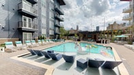 This dynamic amenity space is defined by its bold, contemporary aesthetic and diverse leisure offerings. The centerpiece is a generously sized outdoor pool deck, complete with a shallow sun shelf fitted with modern lounge chairs, traditional poolside seating in teal and white, and views of a prominent city skyline framed by a pergola structure. The surrounding multi-story building features private balconies and large windows overlooking the outdoor space.

Inside, the interior amenity areas showcase an industrial-chic design language, with striking blue-painted exposed ceilings, structural ductwork, and polished tile flooring in a wood-look finish. A comfortable lounge area anchors the interior, featuring plush sectional seating, a large wall-mounted flat-screen television, and distinctive teal pendant lighting. Adjacent to the lounge, a wet bar area is outfitted with stainless steel refrigeration units, dark countertops, and a vibrant teal mosaic tile backsplash, accompanied by bold large-scale artwork on the walls.

A dedicated billiards room rounds out the space, featuring a teal-felt pool table, striped navy and white curtains, and glass doors that allow natural light to filter through, creating an energetic yet inviting atmosphere throughout.