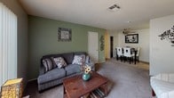 This single-floor space showcases an open-concept layout that seamlessly connects the living and dining areas, creating a cohesive and functional flow throughout. The living area features plush wall-to-wall carpeting in a neutral tone, providing a comfortable foundation for the space. A sage green accent wall adds a warm, inviting pop of color, complementing the neutral beige walls found throughout the rest of the unit. The textured popcorn ceiling adds dimension overhead, while a ceiling-mounted HVAC vent ensures consistent climate control.

The dining area is positioned adjacent to the living space, separated naturally by the transition in wall color from green to yellow. A ceiling fan with a light fixture overhead serves both functional and aesthetic purposes in this zone. Built-in shelving is visible in the background, offering practical storage options.

A door in the living area provides access to additional spaces within the unit. The overall ambiance is cozy and approachable, with warm lighting and a residential character that makes the space feel comfortable and livable. The open sightlines between areas enhance the sense of spaciousness throughout this single-level home.