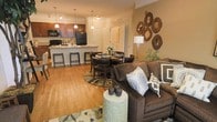 This single-floor residence offers an inviting open-concept layout that effortlessly unites the living, dining, and kitchen areas into one cohesive and functional living space. The living room is anchored by a plush, earth-toned sofa accented with decorative throw pillows, complemented by a stylish ceramic accent table. A gallery-style wall arrangement of circular wood-framed mirrors adds a distinctive artistic touch to the warm, neutral-toned walls. The dining area sits centrally between the living space and kitchen, featuring a round dining table with upholstered seating set atop a patterned area rug, creating an intimate yet accessible setting for meals. The kitchen is well-appointed with rich wood cabinetry, a light-toned countertop island with bar stool seating, black appliances including a refrigerator and microwave, and pendant lighting that adds both function and style. Track lighting runs throughout the ceiling, ensuring the entire open space is well-lit. Warm hardwood-style flooring flows continuously across the living and dining areas, tying the space together visually. The overall ambiance is cozy and well-designed, balancing practical amenities with tasteful décor for comfortable everyday living.