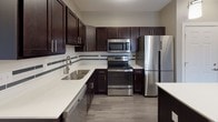 Located at 408 Mill St in Buffalo, NY, this well-designed single-floor residence offers a thoughtfully laid-out interior with modern finishes throughout. The kitchen is a standout feature, showcasing rich dark espresso cabinetry, crisp white quartz countertops, a subway tile backsplash with decorative accent stripe, and a full suite of stainless steel appliances including a refrigerator, range, microwave, and dishwasher. A kitchen island with pendant lighting above adds both function and style, while wood-look flooring ties the space together seamlessly. The open living area is generously proportioned, featuring neutral gray walls, plush carpeting, and a sliding glass door that invites natural light and provides outdoor access. The bedrooms are comfortably sized with soft carpet underfoot, neutral toned walls, and large windows fitted with blinds that allow for ample daylight. The bathroom is well-appointed with dark cabinetry, a wide vanity with white countertop, a large mirror with vanity lighting, a full soaking tub and shower combination with subway tile surround, and grab bars for added accessibility. Throughout the unit, consistent design choices—including brushed nickel hardware and a cohesive neutral color palette—create a clean, modern ambiance.