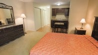 This single-floor studio unit offers an efficient, open-concept layout that combines living, sleeping, and dining functions within one cohesive space. The sleeping area is carpeted in a neutral tone, providing a comfortable foundation, while the remainder of the unit transitions to easy-care hardwood-style flooring. A compact kitchenette is outfitted with dark espresso cabinetry, a granite-style countertop, and a sink, offering essential cooking and prep amenities. Directly adjacent, a small dining area features a dark-toned table and chairs, making practical use of the available space. Two sliding-door closets along one wall provide ample storage, a notable feature for a studio layout. Overhead flush-mount lighting illuminates the unit, supplemented by a floor lamp near the sleeping area. The neutral wall tones throughout contribute to a clean, understated aesthetic. The open arrangement allows flexibility in furniture placement, making the space adaptable to individual needs. Overall, the unit is a functional, well-organized studio designed to maximize utility within a modest footprint.