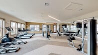 This fitness center in Bryan, Texas, offers a spacious and thoughtfully designed workout environment spread across a single floor. The layout is open and organized, with distinct zones designated for different types of exercise, all united by a cohesive neutral color palette of warm beige and gray tones. Patterned carpet tiles in varying shades of gray add visual interest underfoot while providing a durable, functional surface throughout.

The cardio zone lines an entire wall of large framed windows that allow generous natural light to fill the space and provide outward views of the surrounding exterior. This area is equipped with multiple treadmills, elliptical machines, and stationary bikes. Overhead, linear pendant lighting and ceiling fans complement the natural light and maintain comfortable airflow.

The strength training area is positioned toward the center and perimeter of the room, featuring a multi-station cable machine, adjustable benches, a full dumbbell rack, and a medicine ball station. Large wall-mounted mirrors expand the sense of space and allow users to monitor their form. A mounted flat-screen television is positioned prominently in the strength area.

A secondary alcove area, defined by a contrasting dark accent wall and recessed lighting, provides additional equipment space and contributes to the overall architectural depth of the facility. A reception-style counter near the entrance adds a welcoming, organized touch.