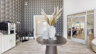 Located at 9015 Kings Grant Dr in Charlotte, NC, this single-floor community clubhouse blends contemporary design with welcoming functionality. The entrance makes an immediate impression with soaring double-height ceilings and a dramatic accent wall clad in a bold three-dimensional geometric wallcovering in deep charcoal tones. A sculptural dark pedestal table anchors the foyer, styled with decorative vases and organic arrangements that set a refined, modern tone.

Light wood-look flooring runs throughout, complementing the crisp white walls and contributing to a bright, airy atmosphere. The main lounge area is generously appointed with plush, cream-toned curved seating chairs, a sleek round coffee table, and curated artwork — including large-scale abstract canvases in gold, black, and grey that add visual depth and sophistication. Floor-to-ceiling windows flood the space with natural light and offer views of the surrounding exterior.

A comfortable seating nook with a neutral sofa and accent pillows provides an additional area for relaxation. The space flows seamlessly toward glass French doors bearing the Conrad branding, which open directly onto an outdoor pool area complete with lounge seating and umbrella-shaded furnishings — extending the living experience outdoors. This thoughtfully designed clubhouse serves as a stylish social hub for the community.