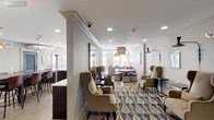 The Club Room at 101 Washington Blvd in Stamford, Connecticut is a thoughtfully designed single-floor amenity space that blends comfort, style, and functionality. The space is organized into several distinct yet interconnected zones, each serving a unique social purpose.

The main lounge area is furnished with a tufted Chesterfield sofa, upholstered armchairs, and a decorative coffee table, all anchored by a striking woven rattan pendant light. Large windows allow generous natural light to fill the room, creating a warm and inviting atmosphere. Colorful artwork adorns the walls, adding a curated, residential feel. A vibrant ikat-patterned area rug defines a secondary seating cluster featuring high-back wing chairs arranged around small side tables, ideal for intimate conversation.

Adjacent to the lounge, a dining and gathering area features a substantial rustic wood high-top table with leather bar stools, illuminated by warm copper pendant lights. A large circular wall art piece serves as a focal point, complementing the industrial-chic aesthetic of this zone.

The kitchenette is fully equipped with stainless steel appliances — including a full-size refrigerator, dishwasher, and microwave — dark espresso cabinetry, a mosaic tile backsplash, and a clean white countertop with an undermount sink, making it well-suited for event hosting and catering needs. Light wood-toned flooring flows throughout, unifying the space cohesively.