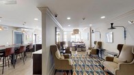 The Club Room at 101 Washington Blvd in Stamford, Connecticut is a thoughtfully designed single-floor amenity space that blends comfort, style, and functionality. The space is organized into several distinct yet interconnected zones, each serving a unique social purpose.

The main lounge area is furnished with a tufted Chesterfield sofa, upholstered armchairs, and a decorative coffee table, all anchored by a striking woven rattan pendant light. Large windows allow generous natural light to fill the room, creating a warm and inviting atmosphere. Colorful artwork adorns the walls, adding a curated, residential feel. A vibrant ikat-patterned area rug defines a secondary seating cluster featuring high-back wing chairs arranged around small side tables, ideal for intimate conversation.

Adjacent to the lounge, a dining and gathering area features a substantial rustic wood high-top table with leather bar stools, illuminated by warm copper pendant lights. A large circular wall art piece serves as a focal point, complementing the industrial-chic aesthetic of this zone.

The kitchenette is fully equipped with stainless steel appliances — including a full-size refrigerator, dishwasher, and microwave — dark espresso cabinetry, a mosaic tile backsplash, and a clean white countertop with an undermount sink, making it well-suited for event hosting and catering needs. Light wood-toned flooring flows throughout, unifying the space cohesively.