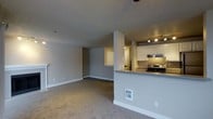 This single-floor space is designed with an open-concept layout that connects the living area and kitchen, creating a seamless flow throughout. The living area is finished with wall-to-wall carpeting in a neutral tone, complemented by light gray walls and white baseboards that contribute to a clean, understated aesthetic. A ceiling fan is positioned overhead, adding to the functional appeal of the space.

The kitchen is situated on one side, partially separated by a half-wall that doubles as a breakfast bar with a gray countertop surface. White cabinetry lines the upper walls, providing ample storage, while the kitchen is equipped with a stainless steel range and an overhead ventilation hood. Track lighting illuminates the kitchen area, ensuring a well-lit workspace.

A hallway visible from the living area leads to additional rooms, suggesting a practical and efficient floor plan. The textured ceiling is consistent throughout, adding subtle visual depth to the space. Overall, the interior presents a neutral, move-in-ready environment with a cohesive color palette and functional features throughout.